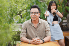 a man wearing glasses sits at a table with his hands folded while a woman stands behind him pointing