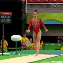 a female gymnast in a red leotard is doing a trick on a balance beam with a omega sign in the background