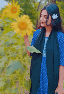 a woman in a blue dress holds a leaf in front of a sunflower