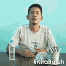 a man is sitting at a table with a bottle of water and a bucket of utensils with the hashtag #kitaboleh