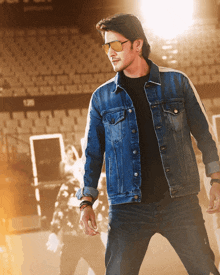 a man wearing a denim jacket and sunglasses stands in front of a empty stadium