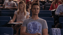 a man wearing a t-shirt that says ' hawaii ' on it sits in a lecture hall with other students