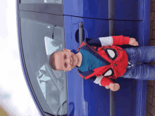 a young boy wearing a spiderman jacket stands next to a blue car