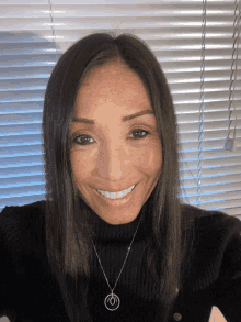 a woman wearing a black sweater and a necklace smiles in front of a window