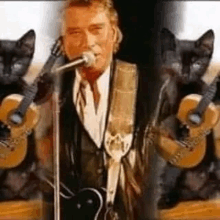 a man in a suit is singing into a microphone while two black cats hold guitars .