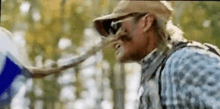 a man wearing a hat and sunglasses is standing in the woods with a snake coming out of his nose .