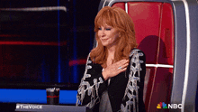 a woman with red hair is sitting in a chair with her hand on her chest in front of a nbc sign