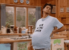 a man is standing in a kitchen wearing a blue shirt that says bun in the oven