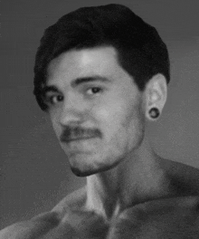 a shirtless man with a mustache and ear plugs looks at the camera