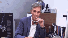 a man in a suit and bow tie is sitting at a desk with a national geographic poster behind him .