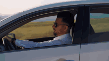 a man in a white shirt and sunglasses is driving a car