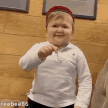 a little boy wearing a hat and a white shirt is standing in front of a wooden wall and smiling .