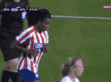 a woman in a red and white striped shirt stands on a soccer field during a game between spa and atl