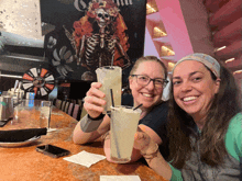 two women toasting with drinks in front of a mural of a skeleton