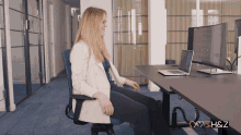 a woman sits at a desk with a laptop and a monitor with the letters h & z below her