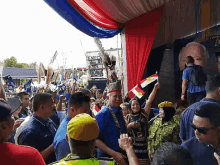 a crowd of people are gathered under a red and blue curtain with a man in a blue shirt holding a flag