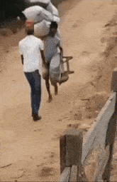 two men are walking down a dirt road and one of them is carrying a large bag on his back .