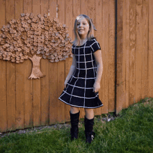 a little girl in a black and white dress stands in front of a wooden fence with a tree carved into it