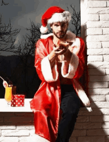 a man in a santa claus costume is sitting on a window sill holding a drink .