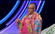 a woman wearing glasses and a colorful jacket is standing in front of a purple background .