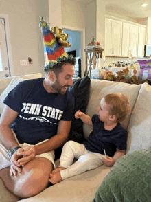 a man in a penn state shirt sits on a couch with a baby