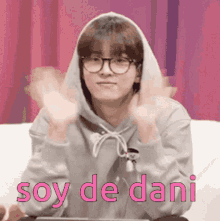 a young man wearing glasses and a hoodie is sitting on a couch with his hands in the air and says soy de dani