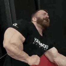 a man with a beard is flexing his muscles in a gym while wearing a black shirt and red shorts .