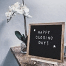 a blackboard with the words `` happy closing day '' written on it is sitting on a table next to a flower .