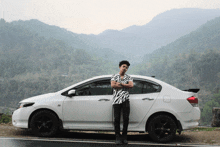 a man stands next to a white car with mountains in the background