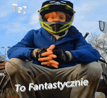 a young boy wearing a helmet and gloves sits with his hands folded in front of a sign that says to fantastycznie