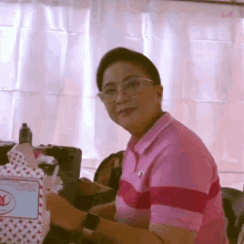 a woman wearing glasses and a pink shirt is sitting at a table in front of a white curtain ..