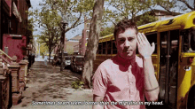 a man in a pink shirt is standing in front of a yellow school bus with his hand on his forehead .