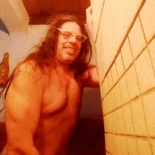 a shirtless man with long hair and glasses leans against a tiled wall