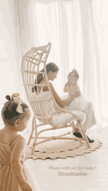 a woman sits in a wicker chair holding a baby while another girl looks on