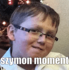 a young boy wearing glasses is smiling in front of a christmas tree and has the words szymon moment written on his face .