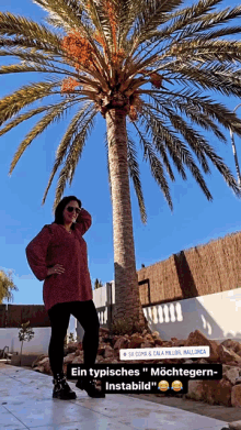 a woman in a red sweater stands in front of a palm tree with a sign that says sa coma & cala millor