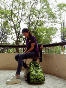 a girl in a purple shirt sits on a chair next to a green backpack that says ' adidas '