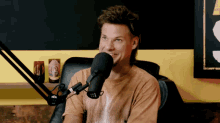 a man in a brown shirt is talking into a microphone with a can of beer on a shelf behind him