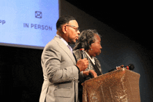 a man and woman stand at a podium in front of a sign that says " in person "