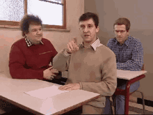 three men are sitting at desks in a room and one man is pointing