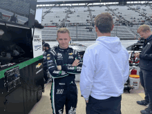 a race car driver wearing a leaf fits uniform talks to a man in a white hoodie