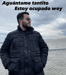 a man wearing sunglasses and a black jacket stands in front of a body of water with the words " aguantame tantito " above him