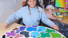 a woman in a blue sweater is sitting at a table with a rainbow on the wall behind her .