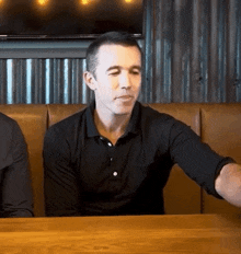 a man in a black shirt is sitting at a table in a restaurant with his arm outstretched .