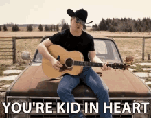 a man sitting on the hood of a truck playing a guitar with the words you 're kid in heart below him