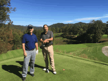 two men stand on top of a golf course with mountains in the background