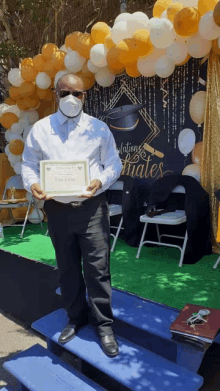 a man wearing a mask is holding a certificate in front of a graduation backdrop .