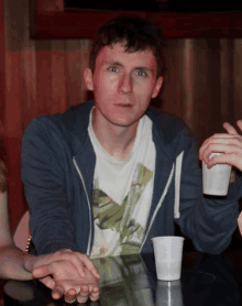 a man in a blue hoodie sits at a table holding a white cup