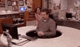 a man is sitting at a desk in front of a computer and a woman is standing behind him .