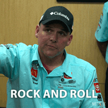 a man wearing a columbia hat and a blue shirt is standing in front of a sign that says rock and roll